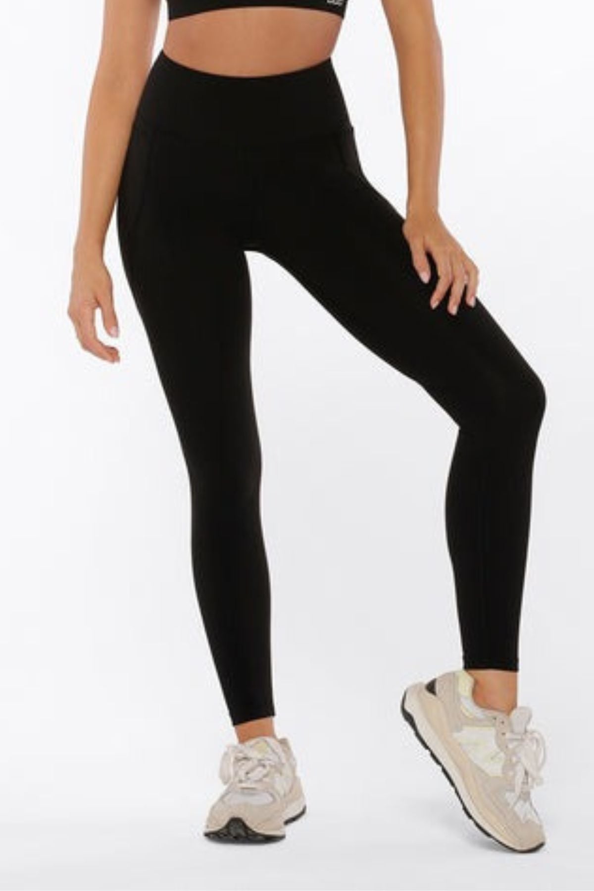A person models the Lorna Jane Amy Phone Pocket Full Length Tech Leggings in black, paired with a matching crop top and light sneakers, standing against a plain white background. The person's head is not shown in the image.