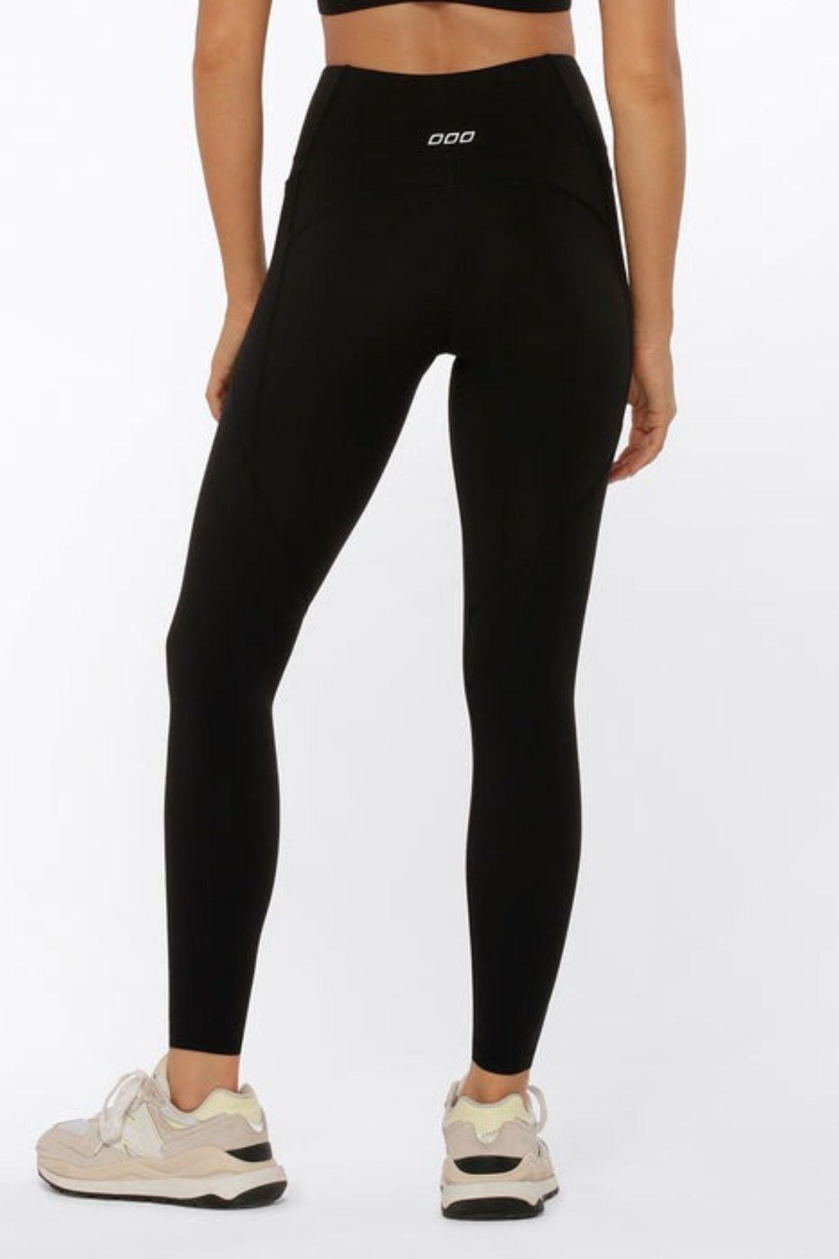 A woman, facing away, wears a black sports bra and Lorna Jane Amy Phone Pocket Full Length Tech Leggings—high-waisted, moisture-wicking—and white sneakers, all against a plain white background.