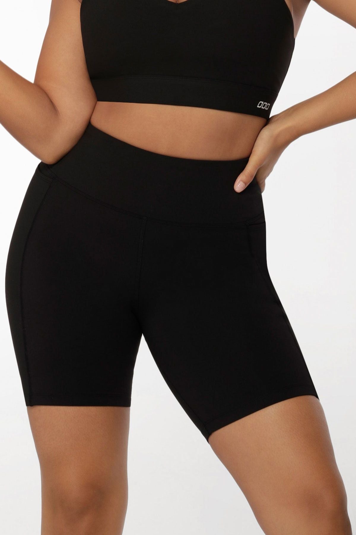 A person models a black sports bra and Lorna Jane Amy Phone Pocket Tech Bike Shorts, featuring a high waist and moisture-wicking fabric, with one hand on their hip against a plain white background.