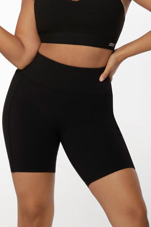 A person models a black sports bra and Lorna Jane Amy Phone Pocket Tech Bike Shorts, featuring a high waist and moisture-wicking fabric, with one hand on their hip against a plain white background.