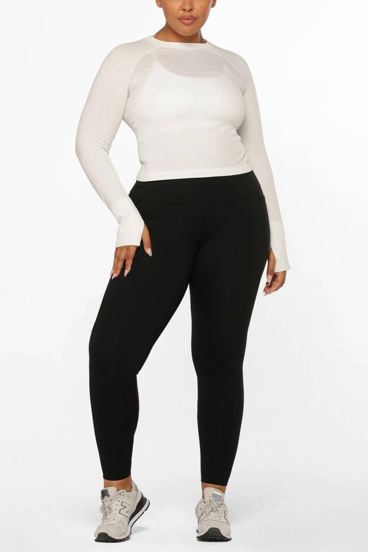 A person stands against a white background in a fitted long-sleeve white top, gray athletic shoes, and Lorna Jane Amy Thermal Phone Pocket Full Length Leggings—black leggings ideal for cold-weather workouts. They pose relaxedly, one hand on their hip.