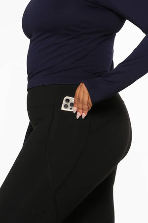 Wearing Lorna Jane Amy Thermal Phone Pocket Full Length Leggings in black, a person places a smartphone into the side pocket of their leggings, dressed in a navy long-sleeve top for cold-weather workouts. Only the lower torso and upper legs are visible.