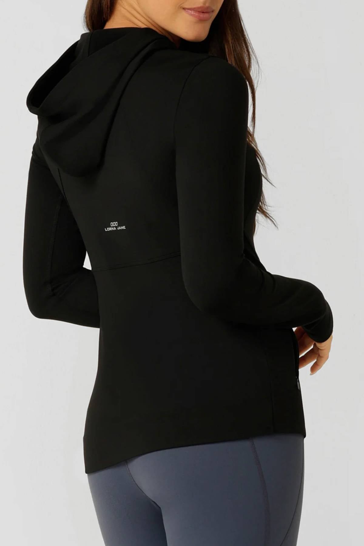 A woman stands facing away, turning slightly to the side, wearing the Lorna Jane Amy Ultimate Thermal Hooded Jacket in black and gray leggings against a plain light background—ideal for winter workouts.