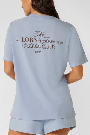 A woman is shown from the back wearing a relaxed fit, light blue Lorna Jane Athletic Club Relaxed T-Shirt and matching shorts. Made from a breathable cotton blend, the tee has "The Lorna Jane Athletic Club EST. 1989" in black script on the back.
