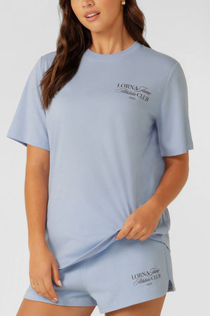 A woman wears the Lorna Jane Athletic Club Relaxed T-Shirt in light blue, featuring "Lorna Jane Athletics Club" print, made from a breathable cotton blend, and stands against a plain white background.