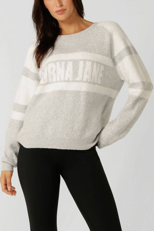 A woman wears the Lorna Jane Cloud Soft Logo Knit Jumper—a light gray and white cotton-blend knit with "LORNA JANE" on the front—paired with black leggings, standing against a plain light background.
