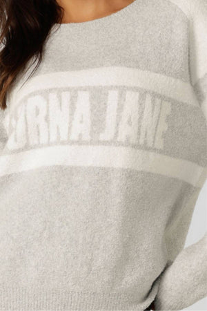 A person wears the Lorna Jane Cloud Soft Logo Knit Jumper, a relaxed mid-length light grey cotton-blend sweatshirt with white stripes and "LORNA JANE" boldly across the chest. Only their upper torso is visible.