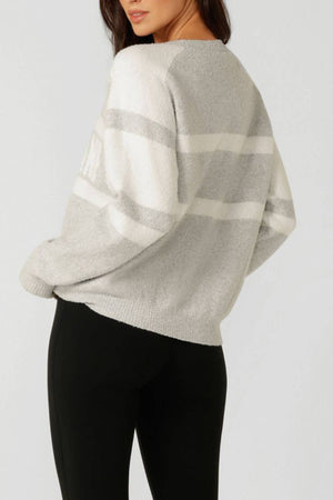 A woman stands sideways in a Lorna Jane Cloud Soft Logo Knit Jumper with light gray and white stripes and black pants. Her head is slightly turned, hair over one shoulder, set against a plain background.