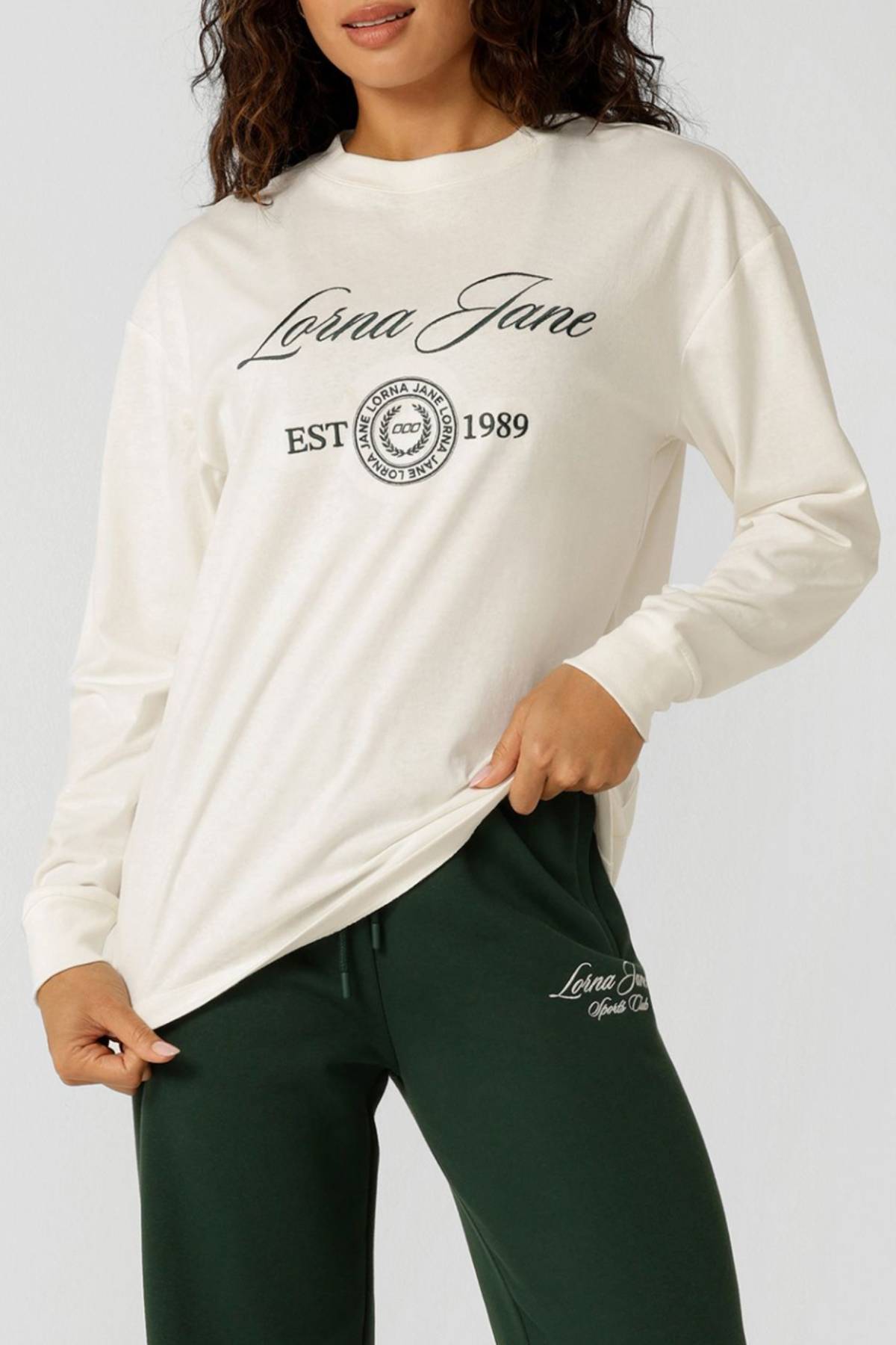 A woman wearing the Lorna Jane Country Club Long Sleeve T-Shirt with "Lorna Jane EST 1989" print and dark green pants poses holding the hem of her shirt.