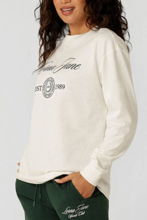Woman in a 100% cotton Lorna Jane Country Club Long Sleeve T-Shirt with "Lorna Jane" and "Est. 1989" text, paired with dark green pants. She has wavy dark hair, one hand in her pocket, and a slight smile.