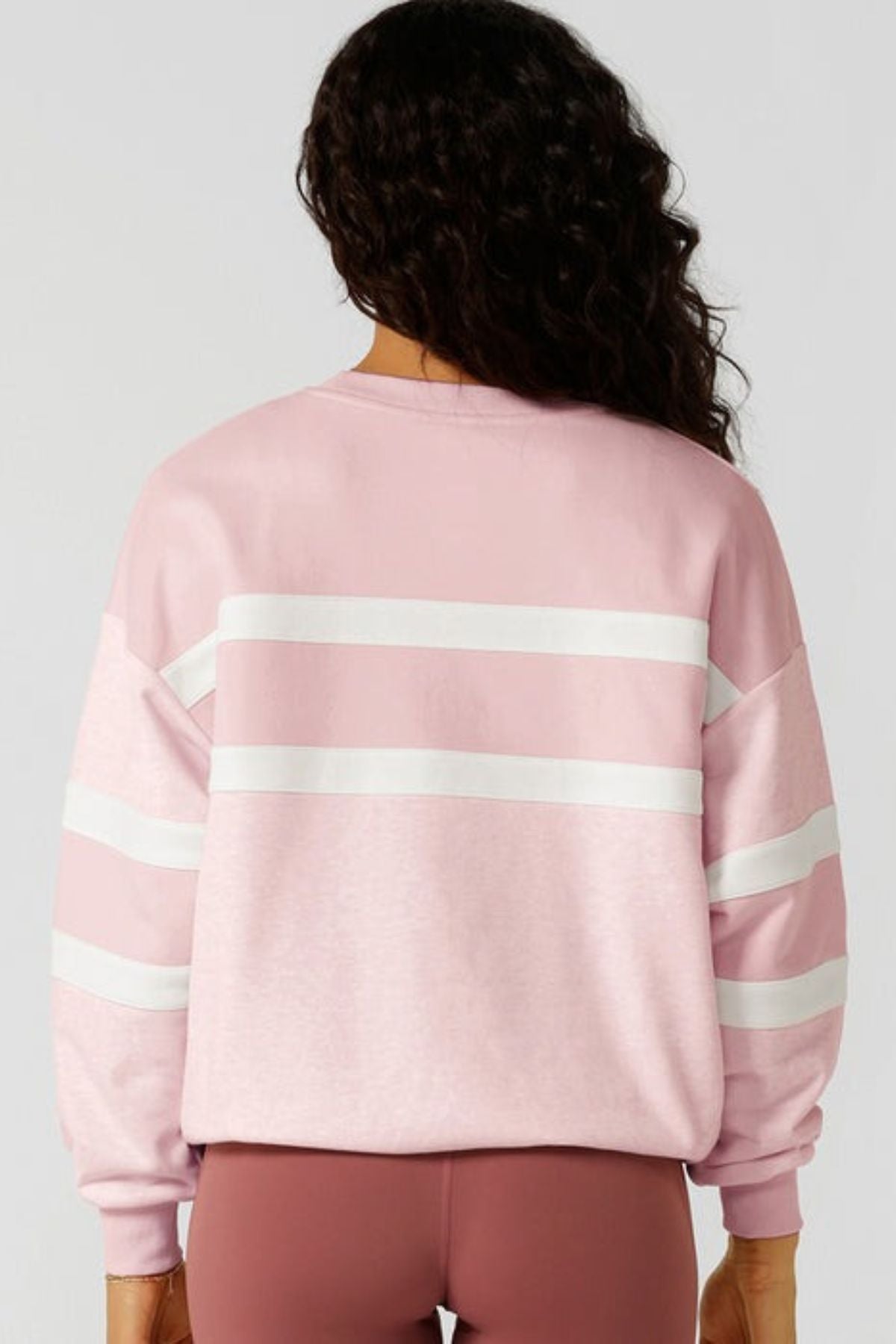 A person with long, wavy dark hair faces away from the camera, wearing a light pink Lorna Jane Dreamer Oversized Sweat with white stripes on the back and sleeves, paired with mauve leggings.