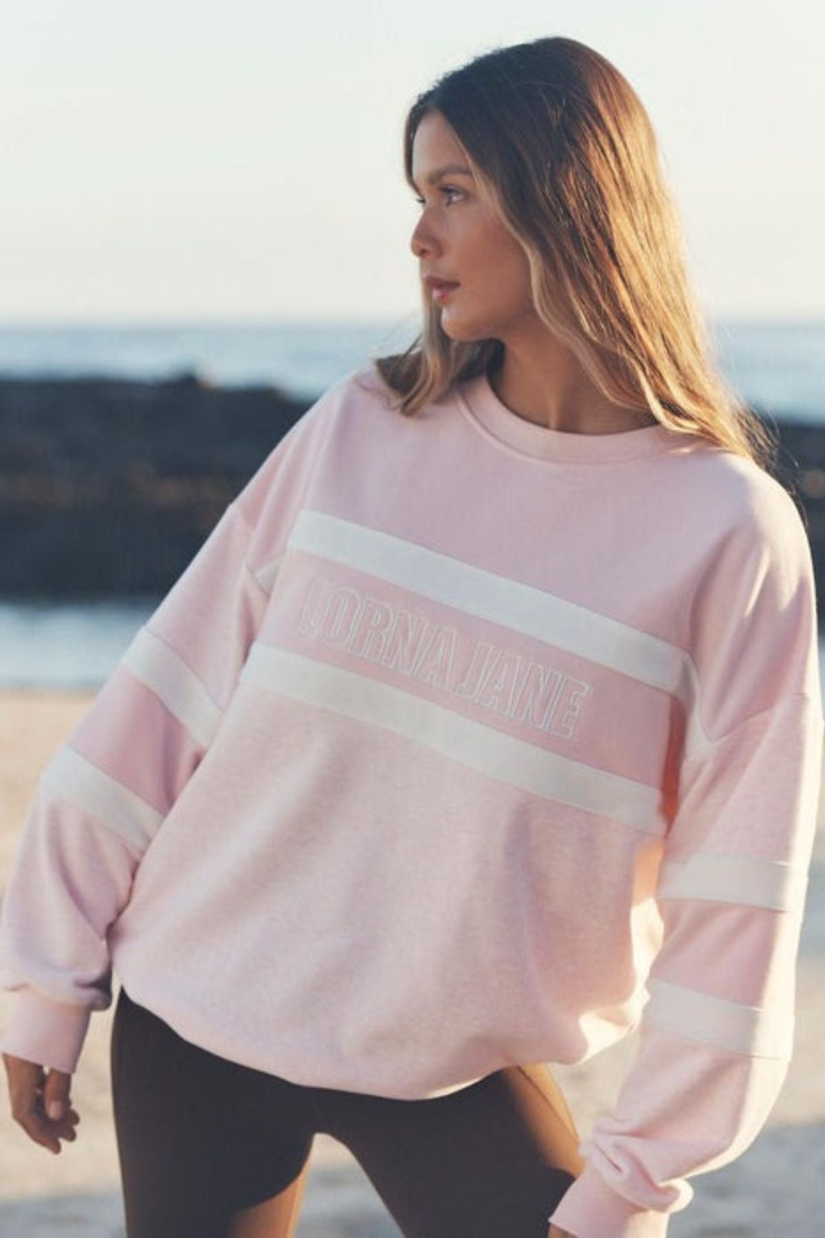 A woman stands on a beach in an oversized fit Lorna Jane Dreamer Oversized Sweat, light pink and white with “BORN JANE” across the front, gazing to the side as the ocean stretches out behind her.