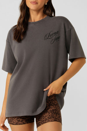 A woman with long, wavy brown hair wears the Lorna Jane Early Riser Washed Boyfriend T-Shirt in dark gray and soft cotton, featuring “Lorna Jane” on the chest, paired with brown leopard print fitted shorts against a neutral background.
