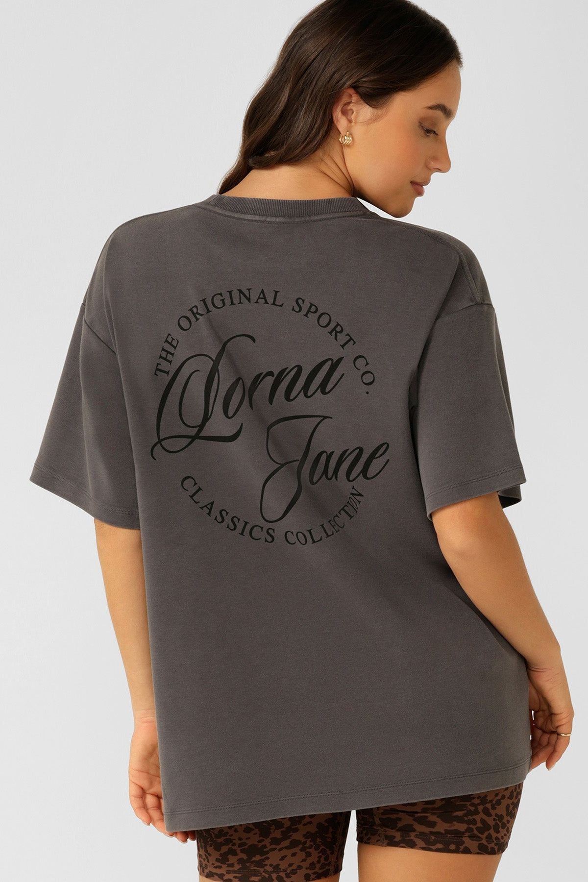 A woman with long brown hair wears the Lorna Jane Early Riser Washed Boyfriend T-Shirt in dark gray, featuring "Lorna Jane" on the back, paired with leopard print shorts against a plain light background.