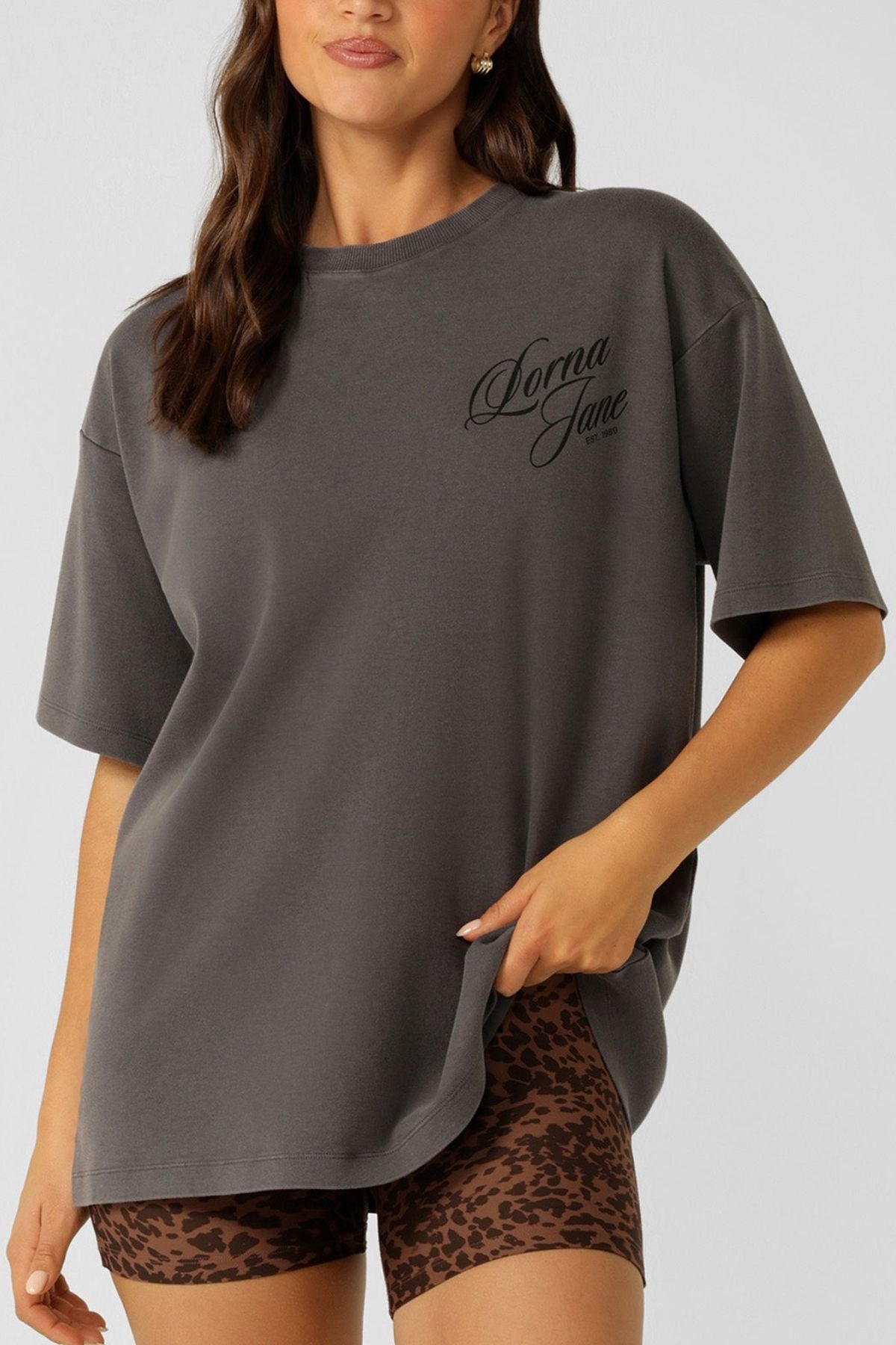 A woman wears the Lorna Jane Early Riser Washed Boyfriend T-Shirt in dark gray with "Donna Jane" text, paired with brown leopard print shorts, standing against a plain light background.