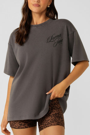 A woman wears the Lorna Jane Early Riser Washed Boyfriend T-Shirt in dark gray with "Donna Jane" text, paired with brown leopard print shorts, standing against a plain light background.