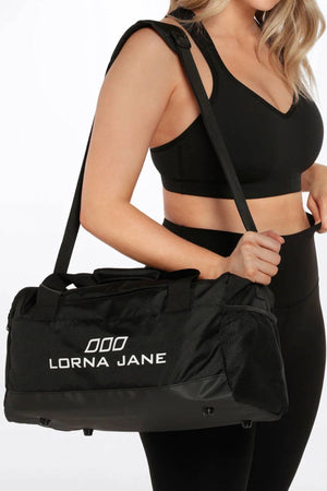 A woman in black athletic wear carries a Lorna Jane Essential Gym Bag with an adjustable shoulder strap, mesh side pockets, a multi-pocket design, and white logo text on the front.