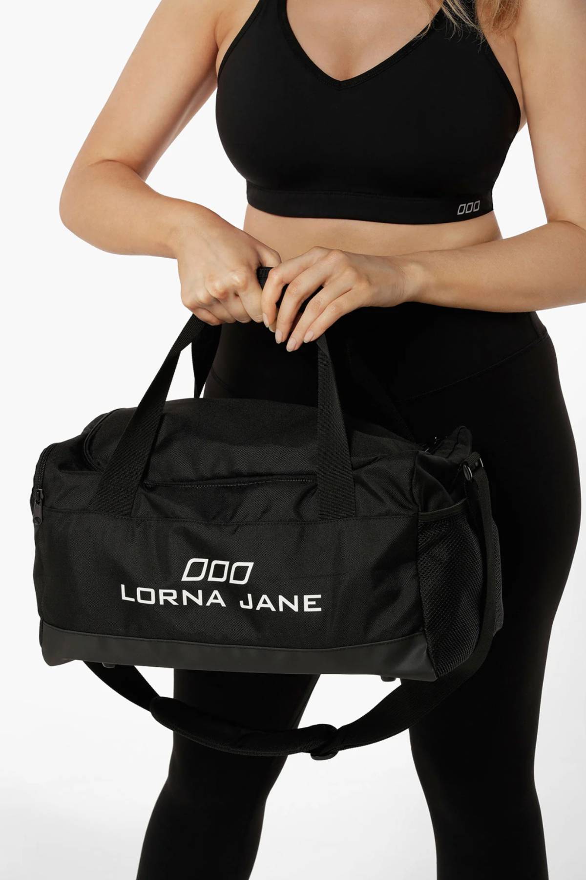 A woman in a black sports bra and leggings holds a Lorna Jane Essential Gym Bag with white lettering, two handles, and an adjustable shoulder strap against a plain white background.