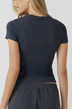 Smiling and showcasing her everyday style against a plain light background, a woman with long blonde hair wears the fitted, short-sleeve Lorna Jane Everyday Rib T-Shirt in a dark hue, paired with matching soft cotton-blend pants and shown from the back.