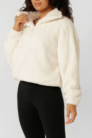 A woman wears the Lorna Jane Everyday Teddy Quarter Zip Sweat, featuring a cozy oversized fit and 100% polyester, with black leggings. She stands against a plain light background, her face partially out of frame.