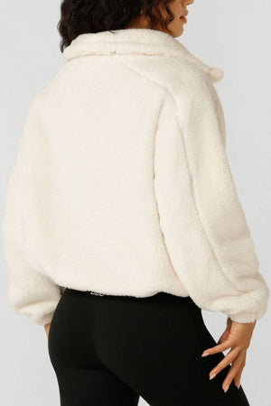 A woman is seen from the back wearing the Lorna Jane Everyday Teddy Quarter Zip Sweat in off-white with an oversized fit and black leggings, standing against a plain background with one hand on her hip.