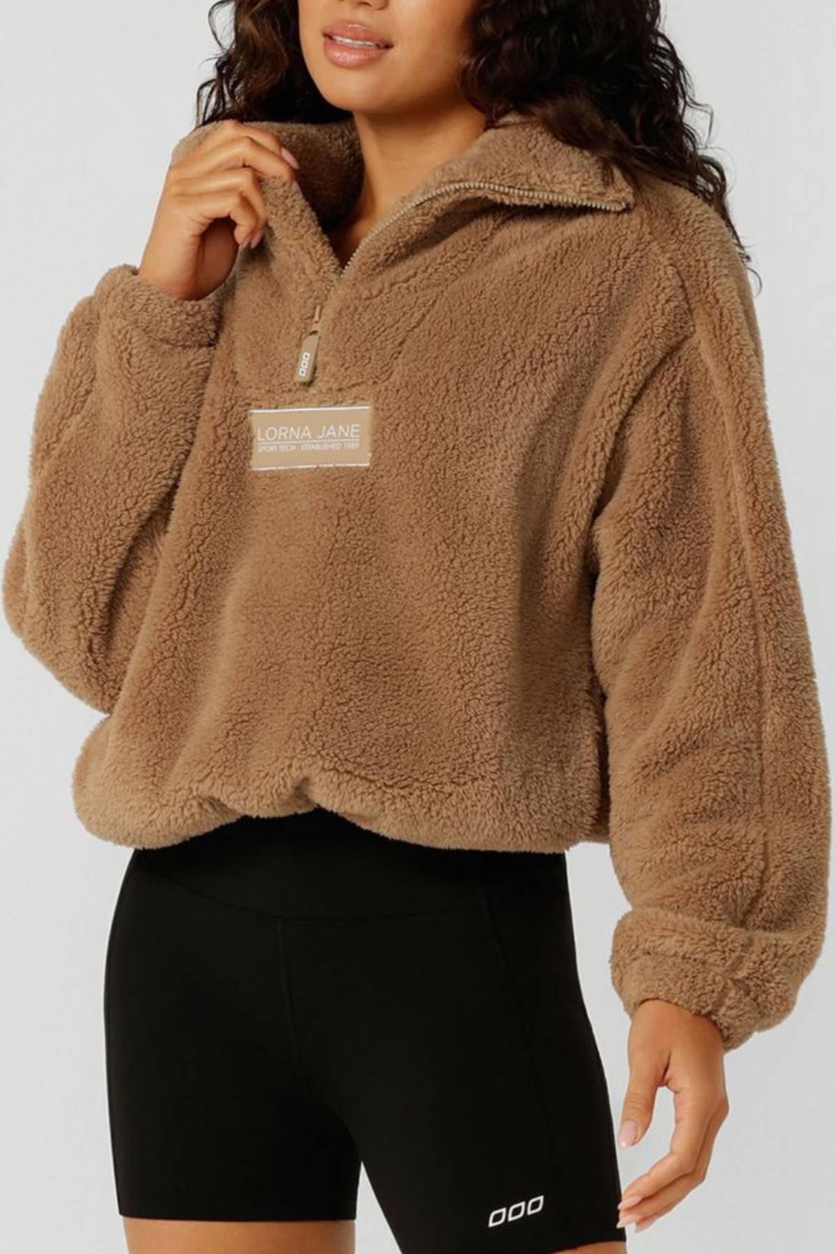 A woman wears a tan Lorna Jane Everyday Teddy Quarter Zip Sweat with black fitted shorts, standing against a plain light background.