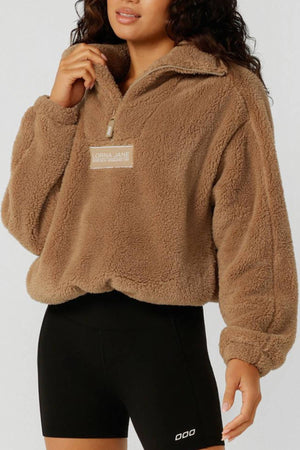 A woman wears a tan Lorna Jane Everyday Teddy Quarter Zip Sweat with black fitted shorts, standing against a plain light background.
