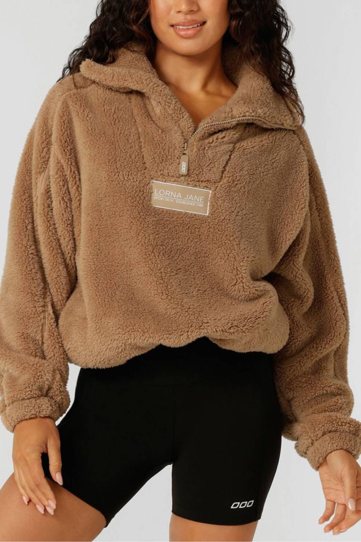 A woman wears the Lorna Jane Everyday Teddy Quarter Zip Sweat, featuring a comfy oversized fit and rubber logo badge, with black athletic shorts as she stands against a plain background.