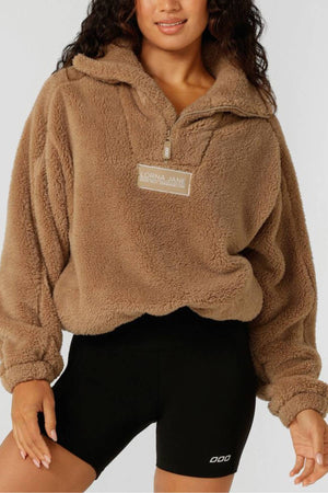 A woman wears the Lorna Jane Everyday Teddy Quarter Zip Sweat, featuring a comfy oversized fit and rubber logo badge, with black athletic shorts as she stands against a plain background.