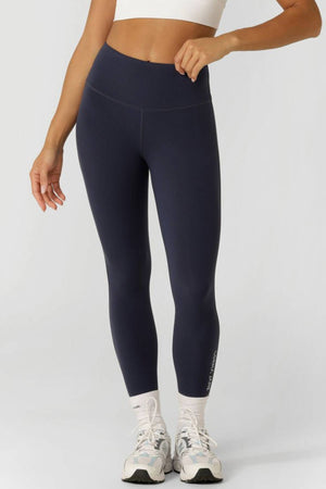 A person wearing a white crop top, navy blue Lorna Jane Lotus No Chafe Phone Pocket Ankle Biter Leggings, white socks, and white athletic shoes stands against a plain light gray background. Their face is not visible.