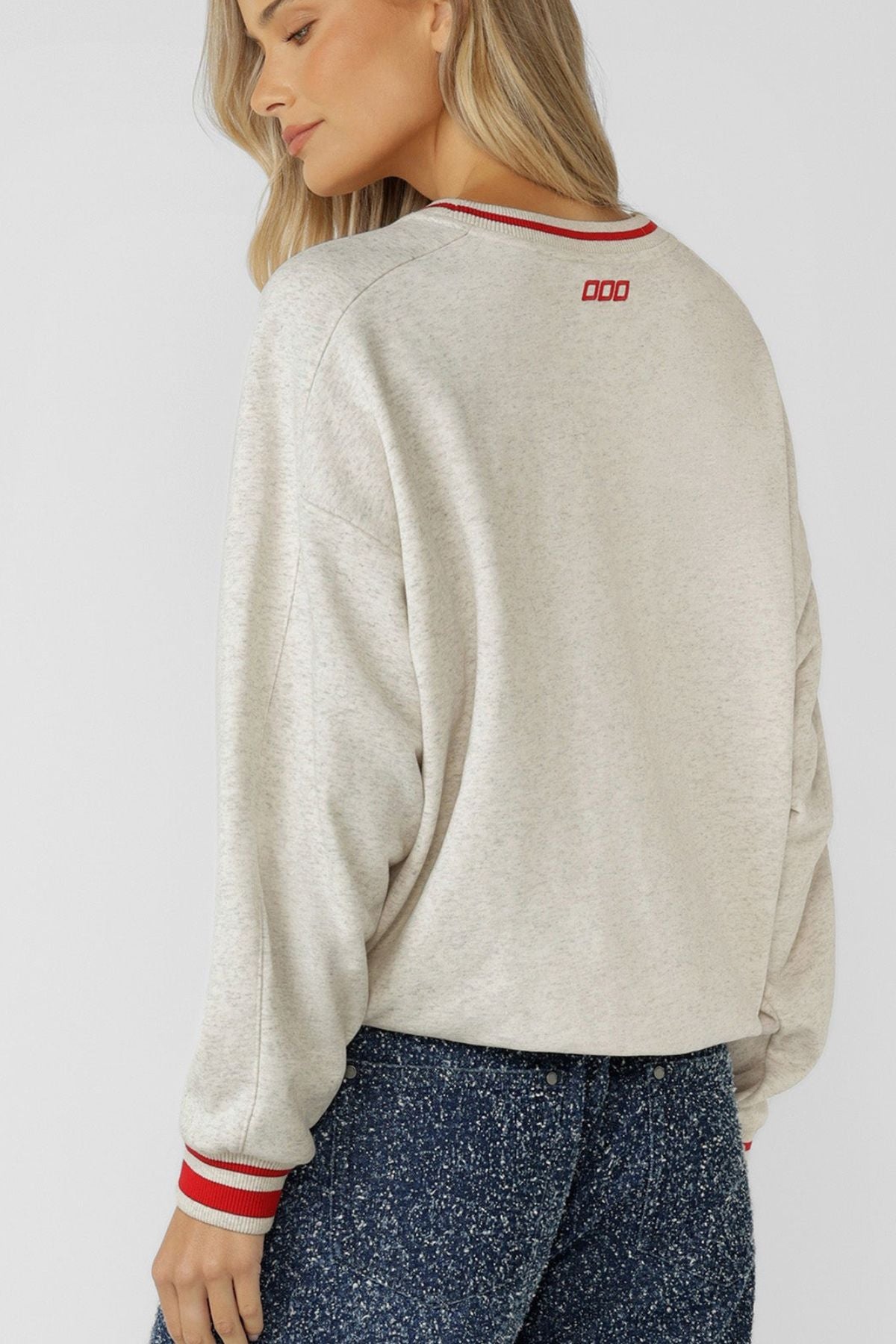 A woman with long blonde hair wears the Lorna Jane Golden Era Sweat, an oversized cotton-blend terry sweatshirt with red and white striped cuffs and collar, paired with blue jeans. She faces away, showing a small embroidered logo near the back neckline.