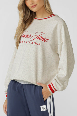 A woman with long, wavy blonde hair wears the Lorna Jane Golden Era Sweat—an oversized cotton blend terry sweatshirt with red striped cuffs and collar, an embroidered "Luxe Athletics" logo—paired with navy athletic pants with white stripes and a logo patch.