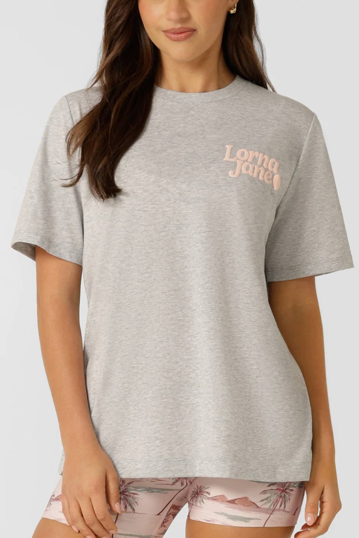 A woman wearing a breathable cotton Lorna Jane Heartbeat Relaxed T-Shirt in light gray and pink palm tree shorts, standing against a plain background.