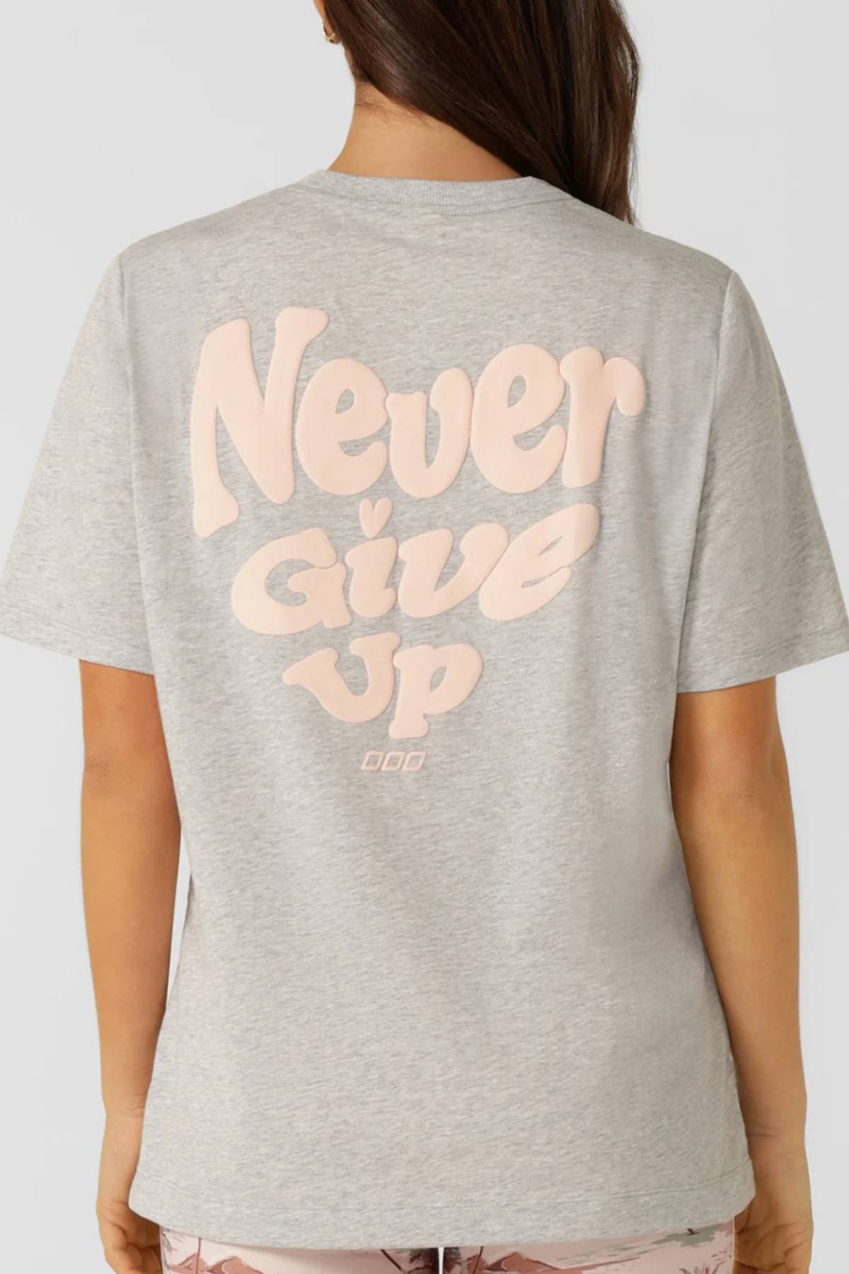 A person with long brown hair is wearing a Lorna Jane Heartbeat Relaxed T-Shirt made from breathable cotton, featuring large, light pink letters on the back that say "Never Give Up.