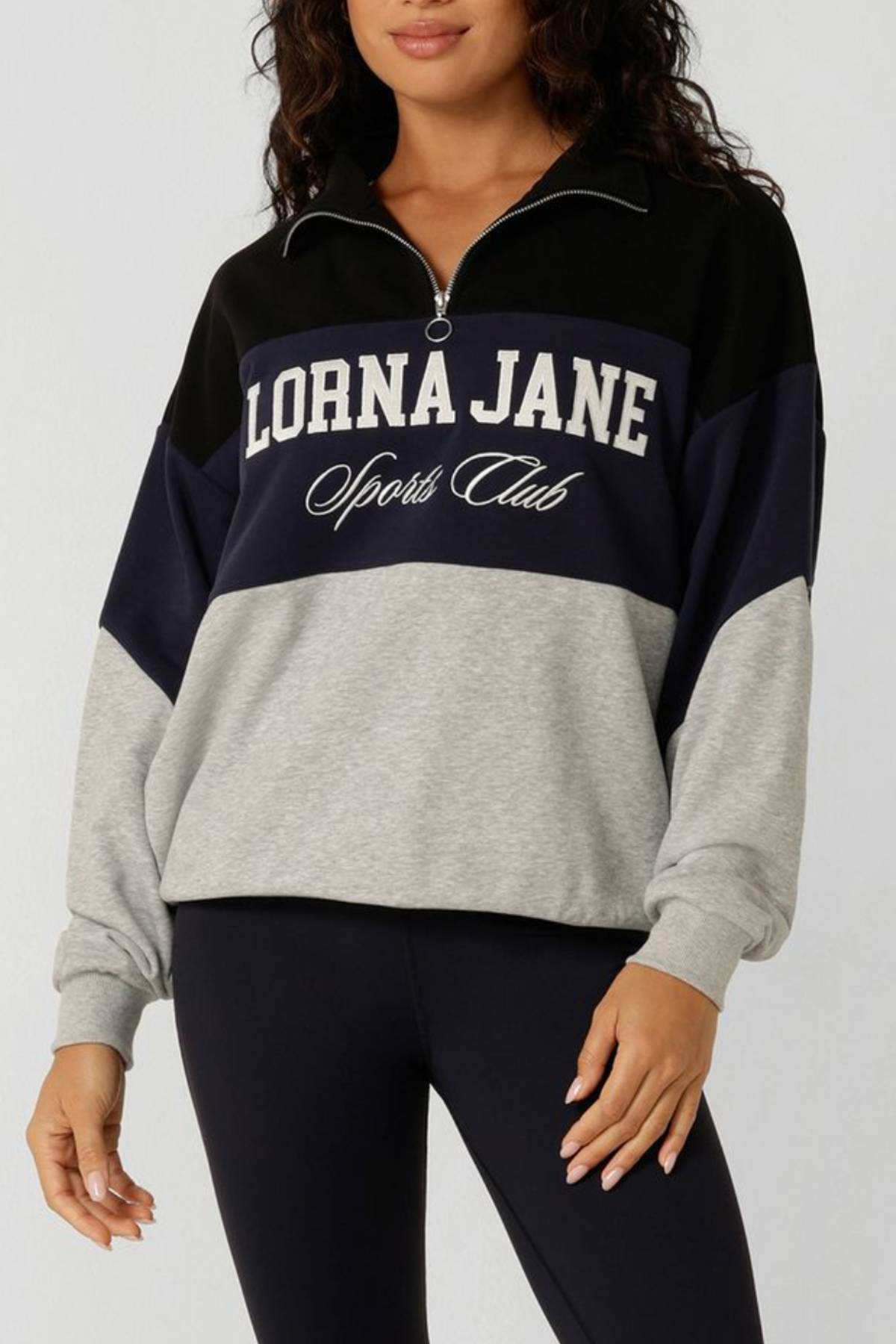 A woman wears the Lorna Jane Heritage Iconic Quarter Zip Sweat in black, navy, and gray with "LORNA JANE Sports Club" on the front, paired with black leggings. Her face is partly out of frame.