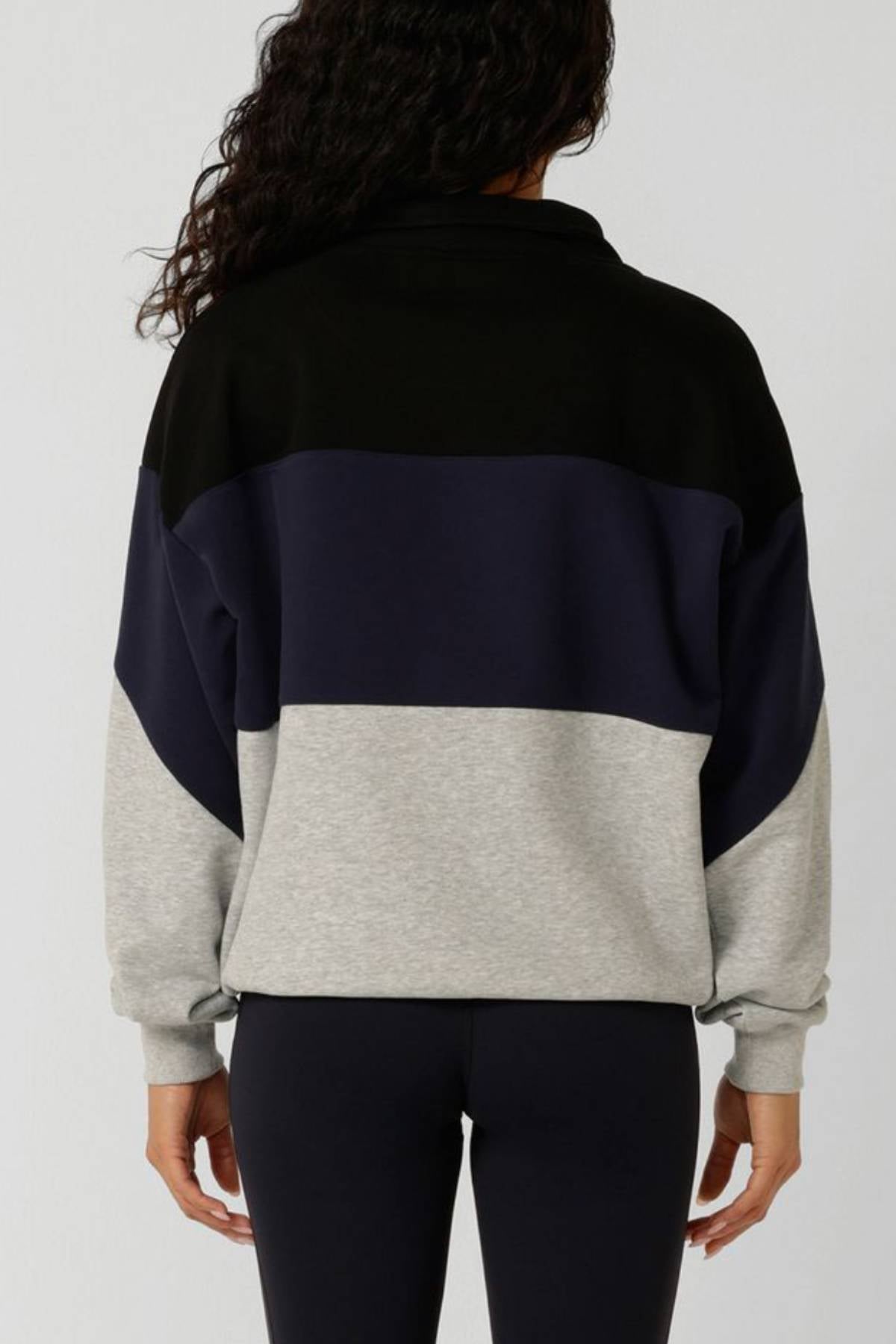 A person with curly dark hair is shown from the back wearing the Lorna Jane Heritage Iconic Quarter Zip Sweat in color-blocked black, navy, and gray, paired with black leggings against a plain light background.