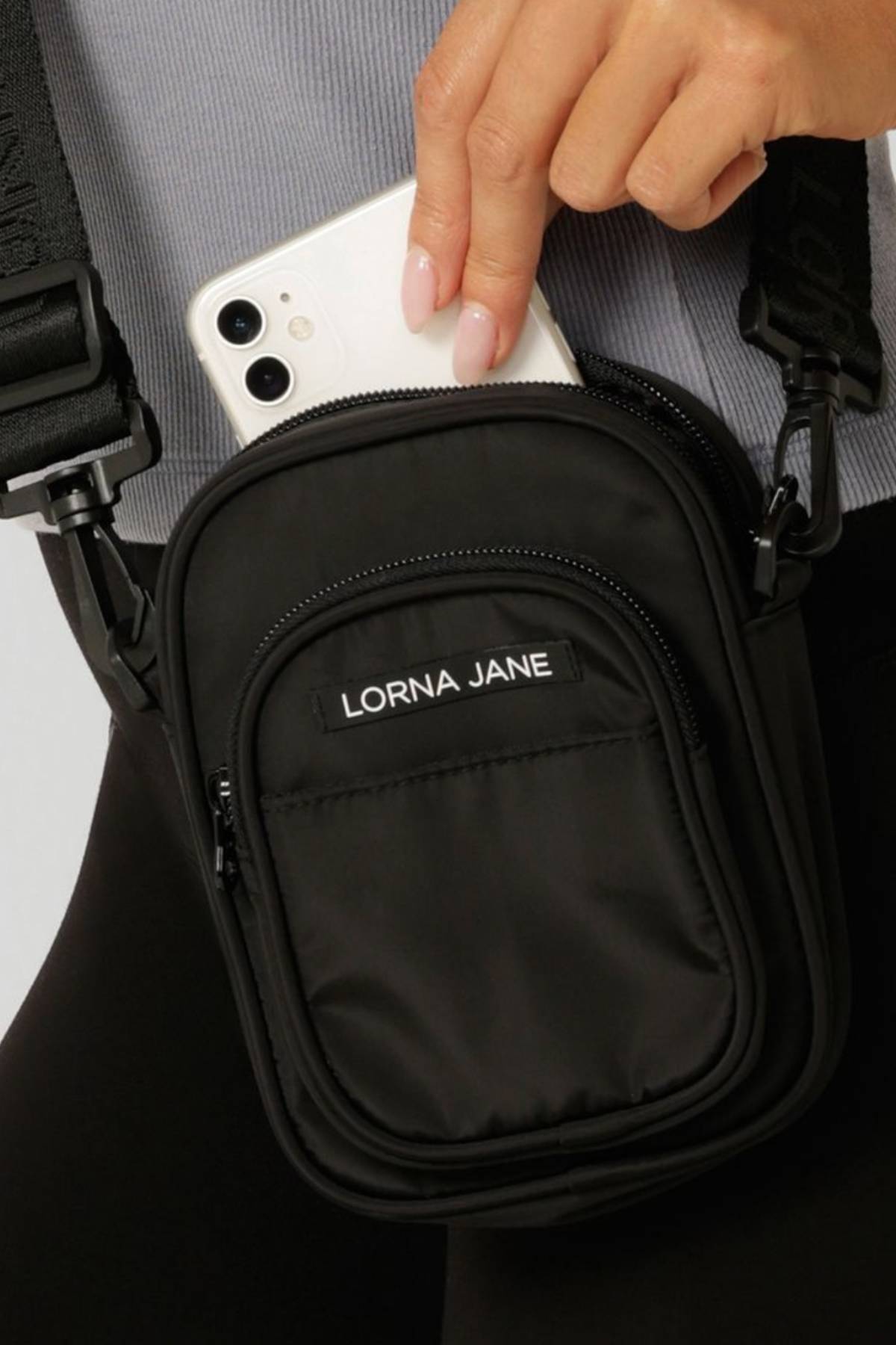 A person puts a white smartphone into a small black Lorna Jane LJ Crossbody Bag featuring a zipper, multiple pockets, and the brand name displayed on the front pocket.