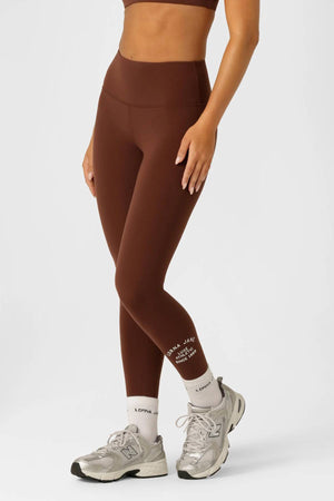 A person in a brown sports bra and Lorna Jane Limited Edition Lotus Full Length Leggings stands with arms crossed, wearing white socks and gray athletic shoes against a plain white background.