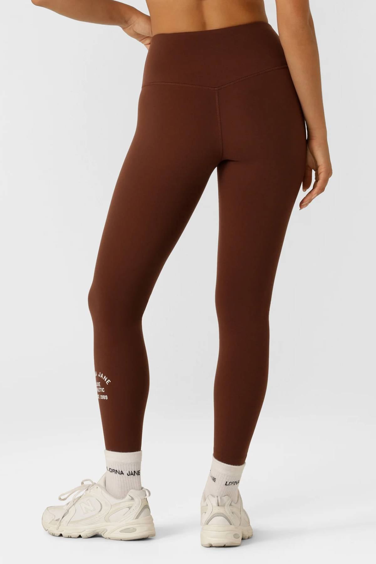 A woman faces away wearing Lorna Jane Limited Edition Lotus Full Length Leggings in brown, paired with white athletic sneakers and "LORNA JANE" socks. The leggings have a small white logo on the lower left leg.