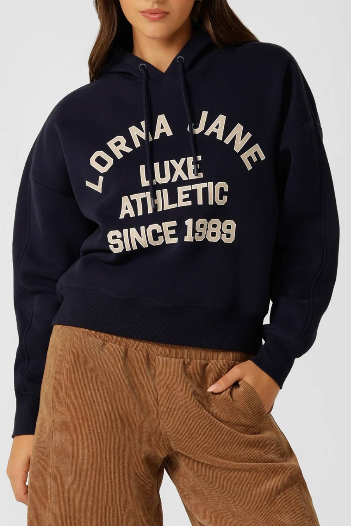 A woman wears the Lorna Jane Limited Edition Lotus Hoodie with "Lorna Jane Luxe Athletic Since 1989" in white, paired with tan corduroy pants. The image, cropped shoulder to mid-thigh, highlights a casual and stylish look.