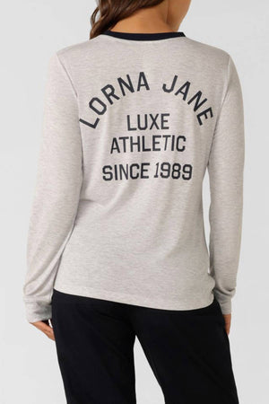Wearing the Lorna Jane Limited Edition Lotus Long Sleeve Top in light grey, featuring bold black "LORNA JANE LUXE ATHLETIC SINCE 1989" text on the back, paired with black pants for a modern, standout look.