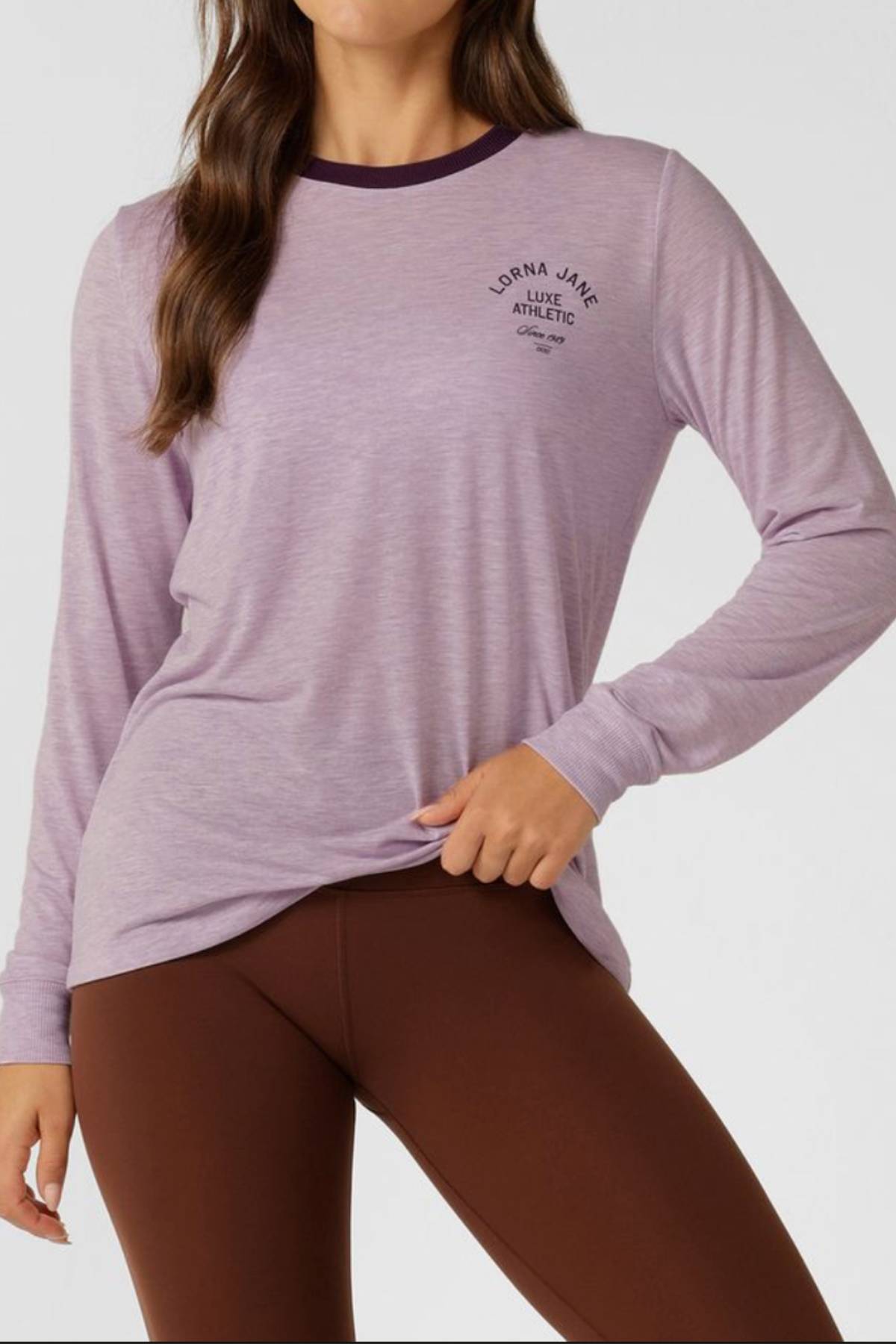 A woman models the Lorna Jane Limited Edition Lotus Long Sleeve Top in light purple with a dark neck trim and small left chest logo, paired with brown leggings. She poses confidently, one hand on her hip.