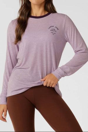 A woman models the Lorna Jane Limited Edition Lotus Long Sleeve Top in light purple with a dark neck trim and small left chest logo, paired with brown leggings. She poses confidently, one hand on her hip.