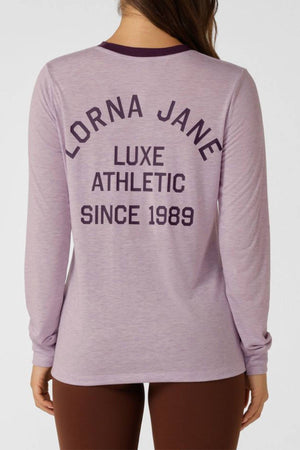 A person faces away from the camera wearing the Lorna Jane Limited Edition Lotus Long Sleeve Top, featuring "LORNA JANE LUXE ATHLETIC SINCE 1989" printed on the back in dark purple letters.