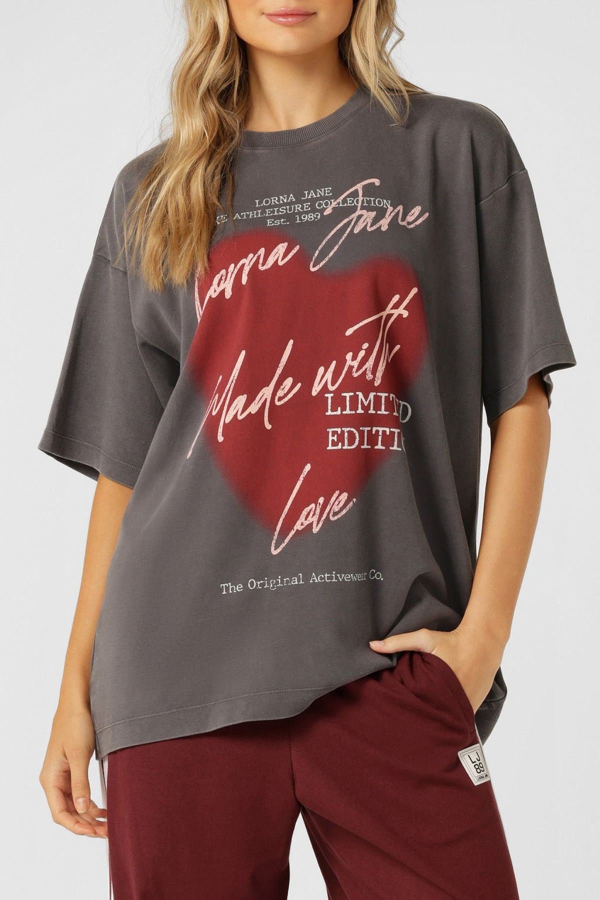 A woman with long, wavy blonde hair wears the Lorna Jane LJ With Love Boyfriend T-Shirt in dark gray—oversized, with red and white cursive text and a red heart—paired with burgundy pants. Her hand is in her pocket.