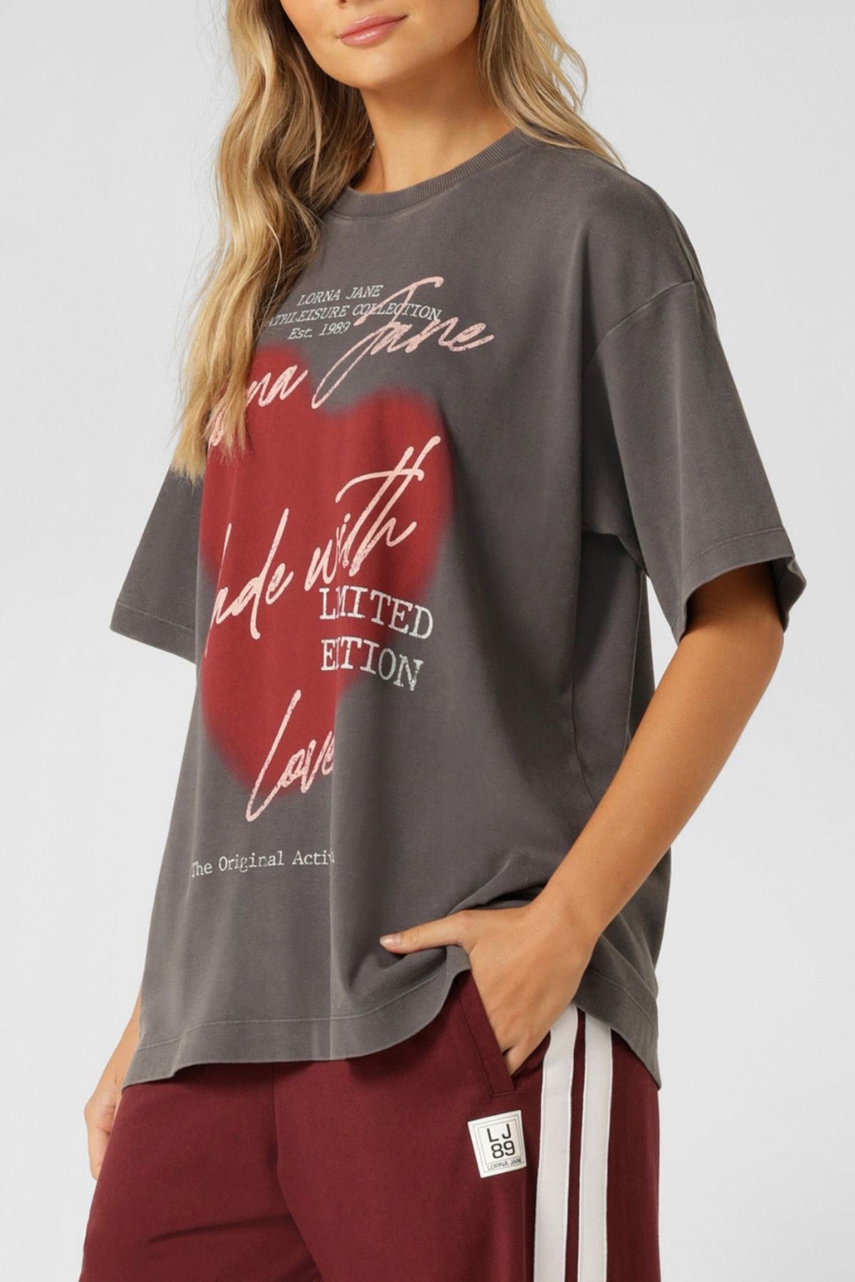 A woman wears the Lorna Jane LJ With Love Boyfriend T-Shirt, an oversized gray tee with red and white graphic text, paired with maroon pants featuring white side stripes and a small patch by the pocket.