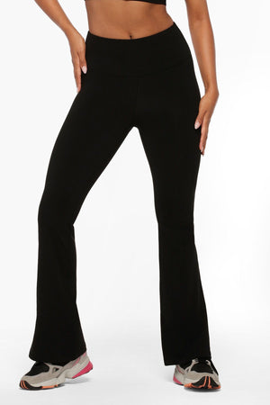 Wearing Lorna Jane Lotus Flared Legging with a high-rise waistband, a black crop top, and gray sneakers with pink accents, a person stands with hands on hips against a white background.