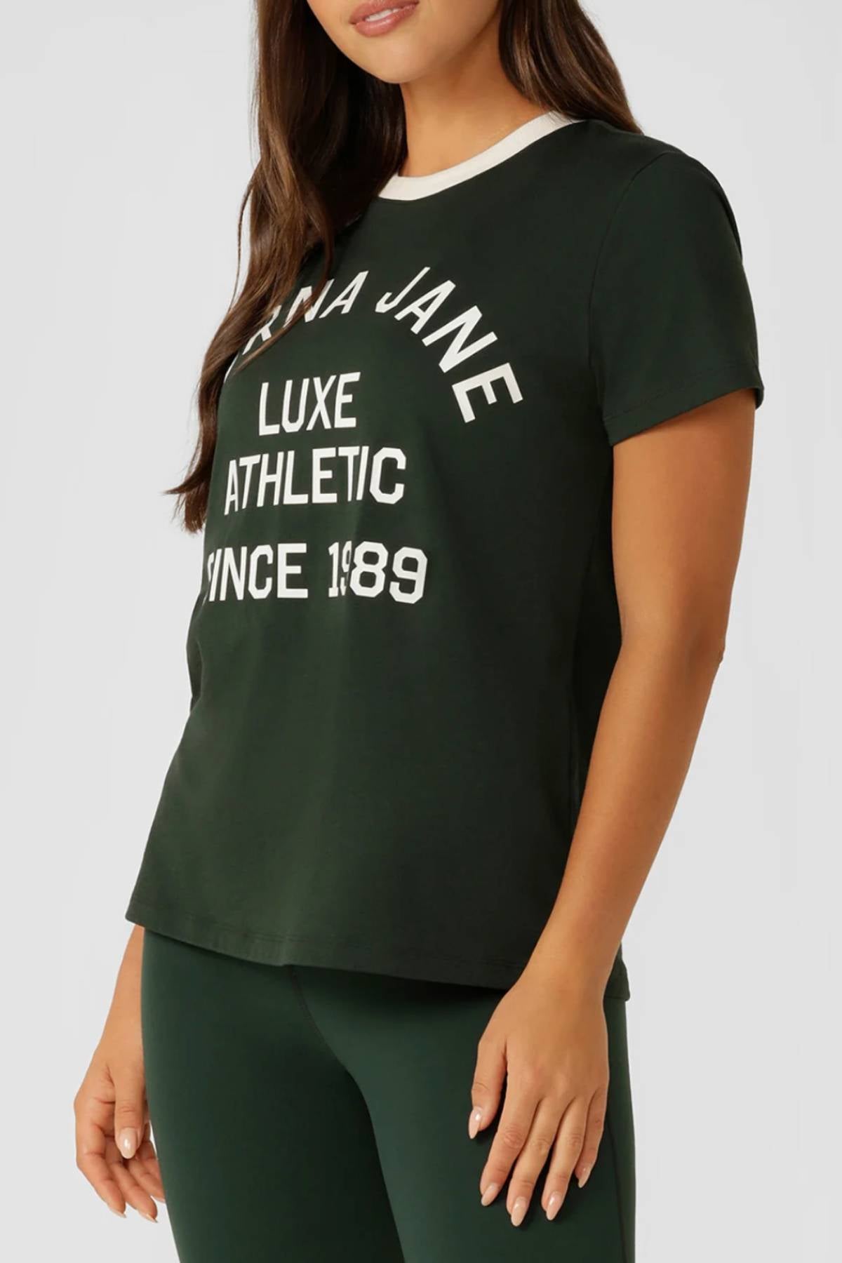 A woman with long brown hair wears the Lorna Jane Lotus Heritage Ringer T-Shirt in dark green with a white collar and "LUXE ATHLETIC SINCE 1989" limited edition logo, paired with matching green leggings.