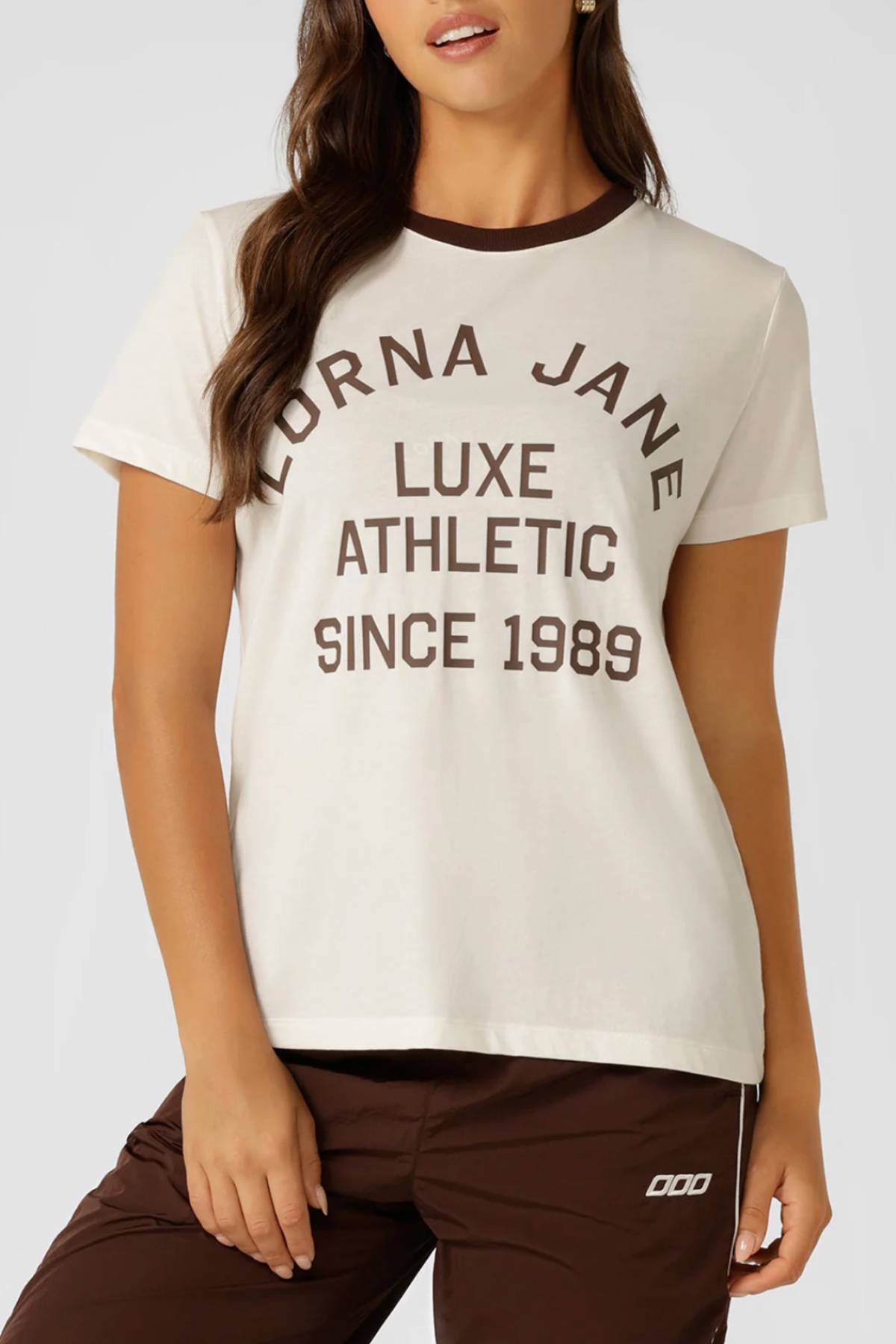 A woman wears the Lorna Jane Lotus Heritage Ringer T-Shirt in cream with dark brown trim and "Lorna Jane Luxe Athletic Since 1989" text, paired with matching brown athletic pants. Only her upper body is visible.