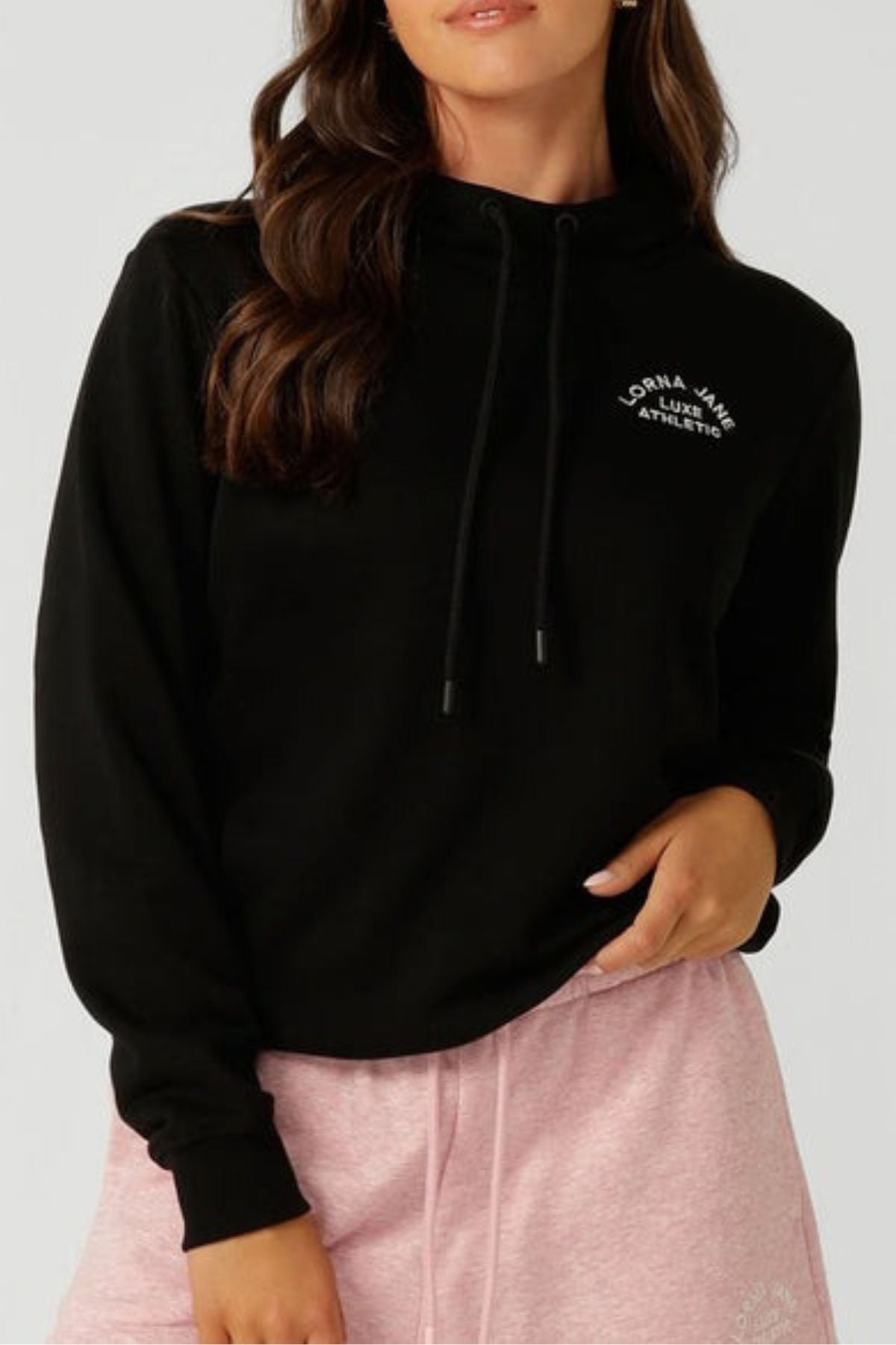 A woman stands with one hand on her hip and the other by her side, wearing the Lorna Jane Lotus Hoodie—a breathable cotton blend hoodie with "Lorna Jane Athletic" on the chest—paired with light pink shorts.